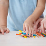 A child's hand reaching for a pile of multi coloured sticks from the PlanToys Mini Pick-Up Sticks set. An adult's hand can be seen on the right reaching for the yellow coloured stick. 
