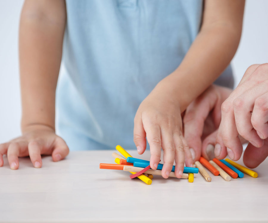 A child's hand reaching for a pile of multi coloured sticks from the PlanToys Mini Pick-Up Sticks set. An adult's hand can be seen on the right reaching for the yellow coloured stick. 
