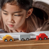 A close up of three vehicles from the PlanToys Mini Cars Set 
laced on a wooden surface. There is a yellow, grey and red car. A child can be seen behind the cars.