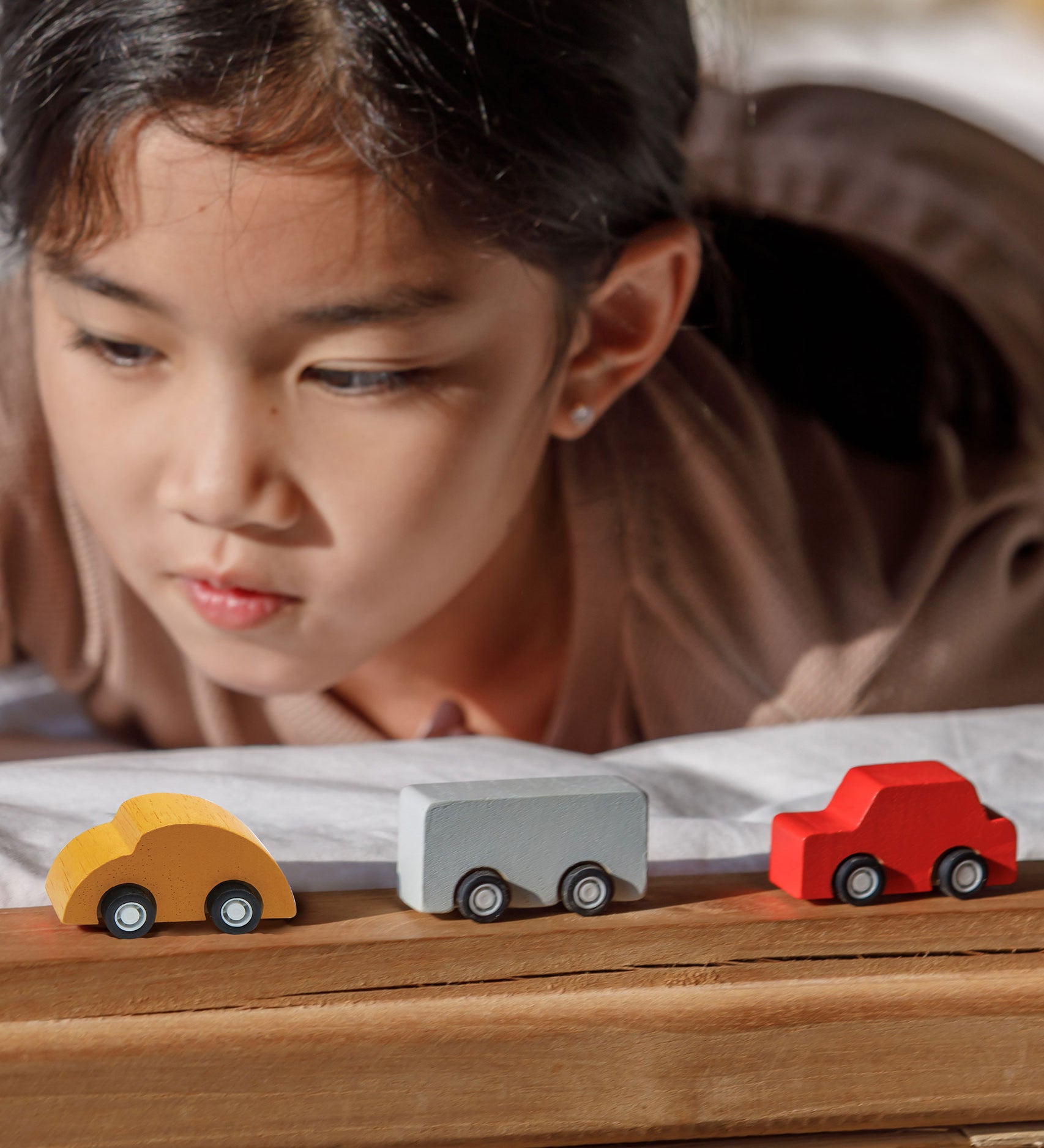 A close up of three vehicles from the PlanToys Mini Cars Set 
laced on a wooden surface. There is a yellow, grey and red car. A child can be seen behind the cars.