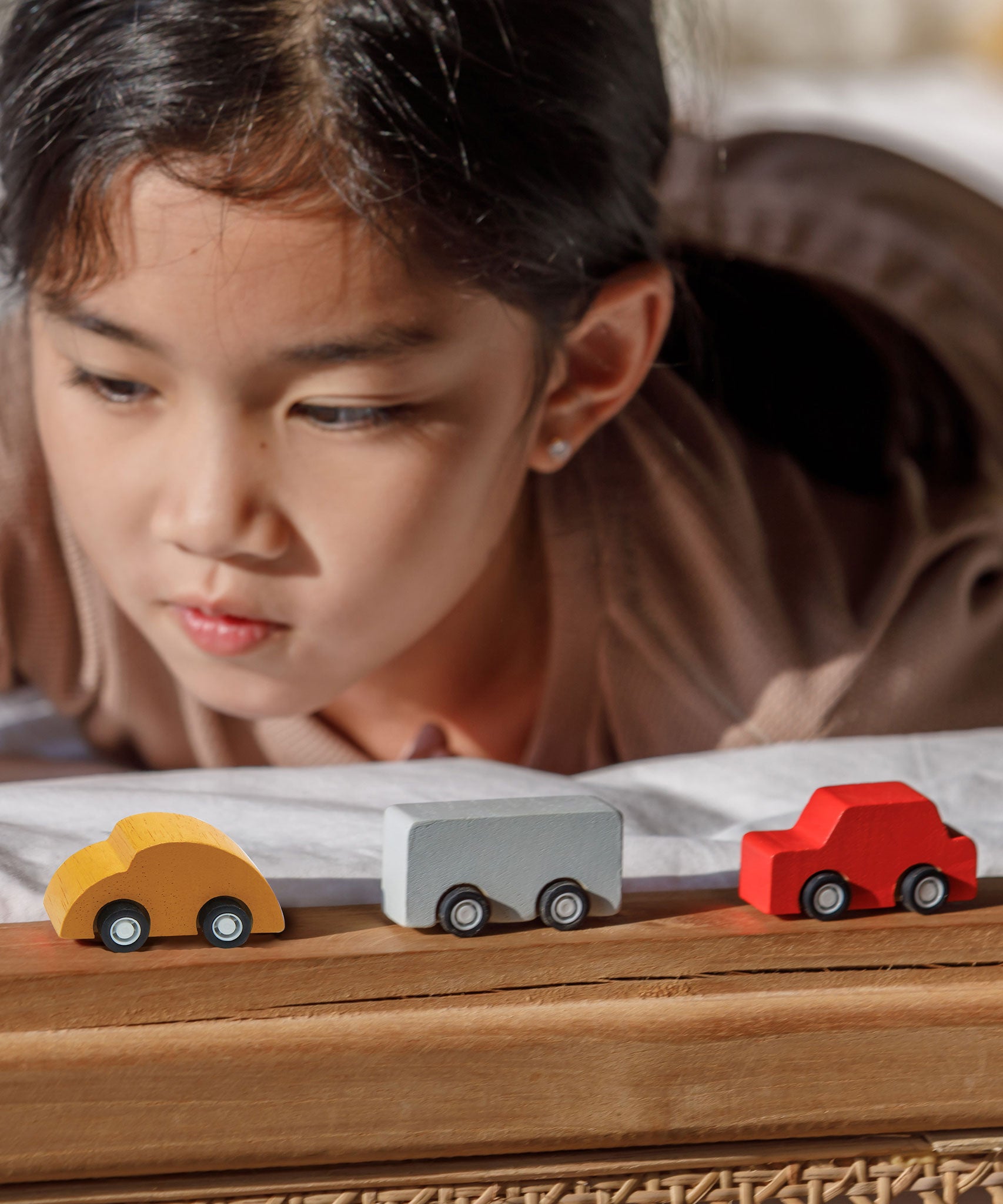 A close up of three vehicles from the PlanToys Mini Cars Set 
laced on a wooden surface. There is a yellow, grey and red car. A child can be seen behind the cars.