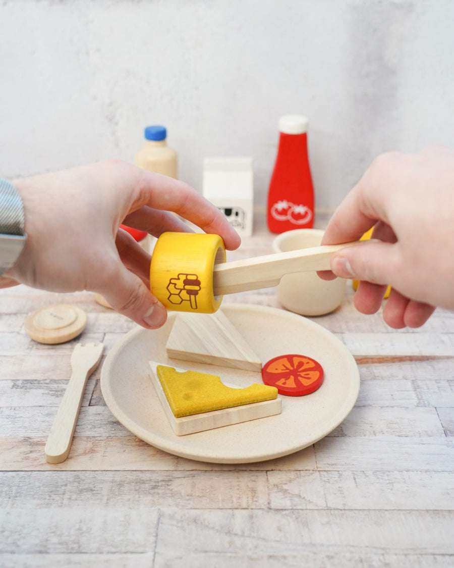 Close up of some hands holding a wooden pot of PlanToys play food honey over some toy slices of bread