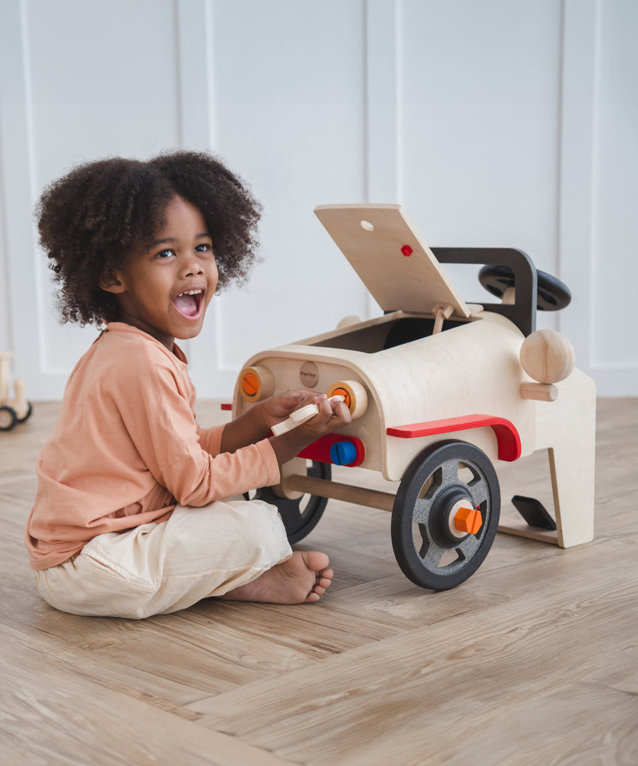 A child playing with the PlanToys Motor Mechanic. The child is tinkering with the headlights using one of the tools and has a big smile on their face.  
