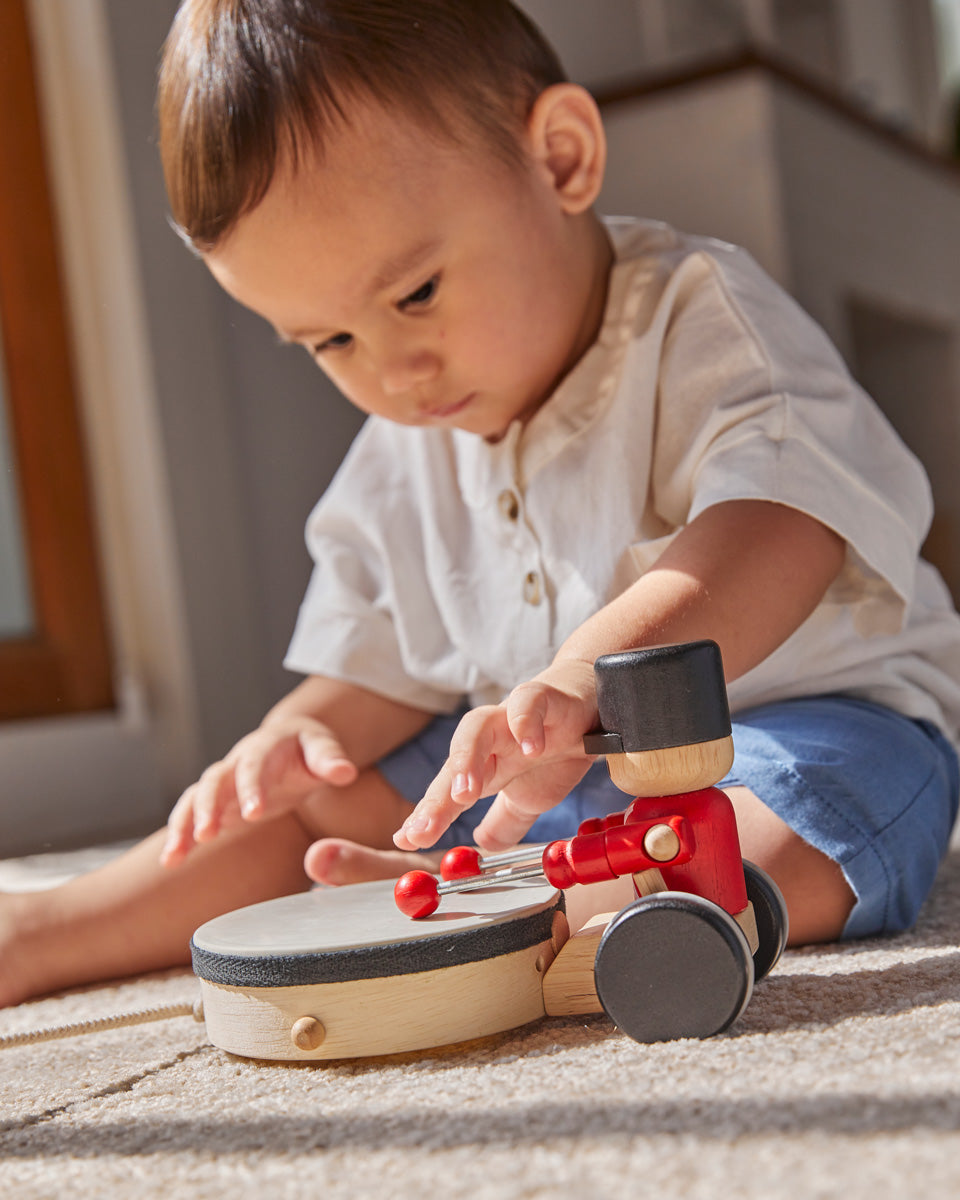 Young boy sat on a carpet playing with the PlanToys wooden pull along drummer