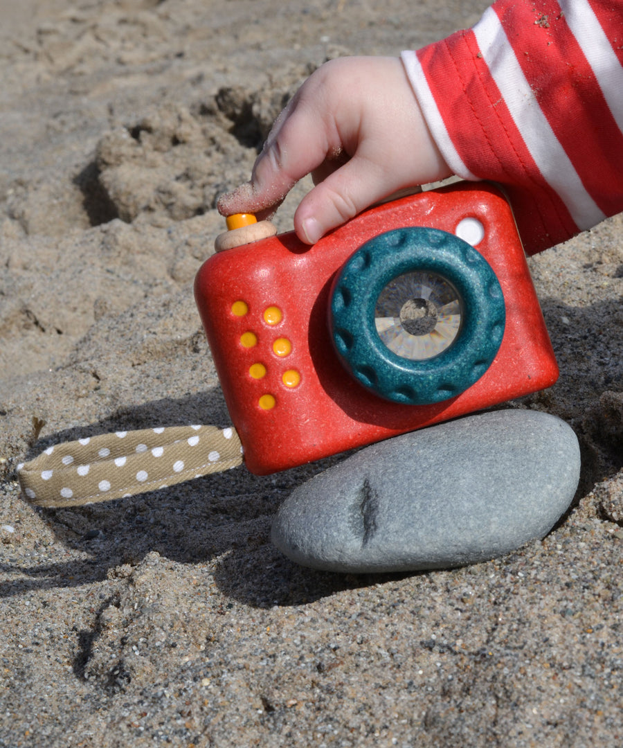 A Plan Toys My First Camera placed on a smooth pebble on the sand. A child's finger can be seen pressing the shutter release button. 