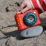 A Plan Toys My First Camera placed on a smooth pebble on the sand. A child's finger can be seen pressing the shutter release button. 