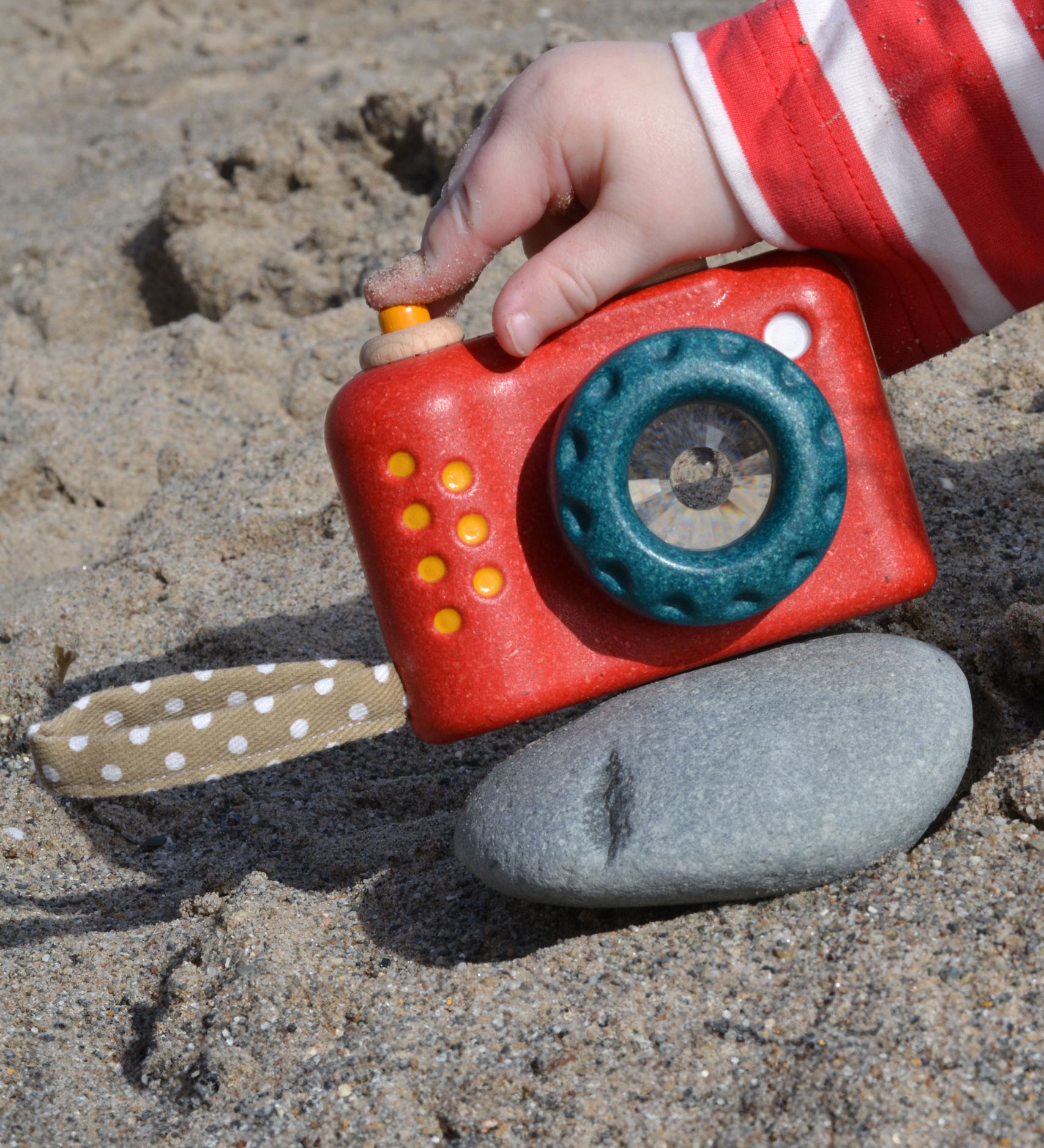 A Plan Toys My First Camera placed on a smooth pebble on the sand. A child's finger can be seen pressing the shutter release button. 