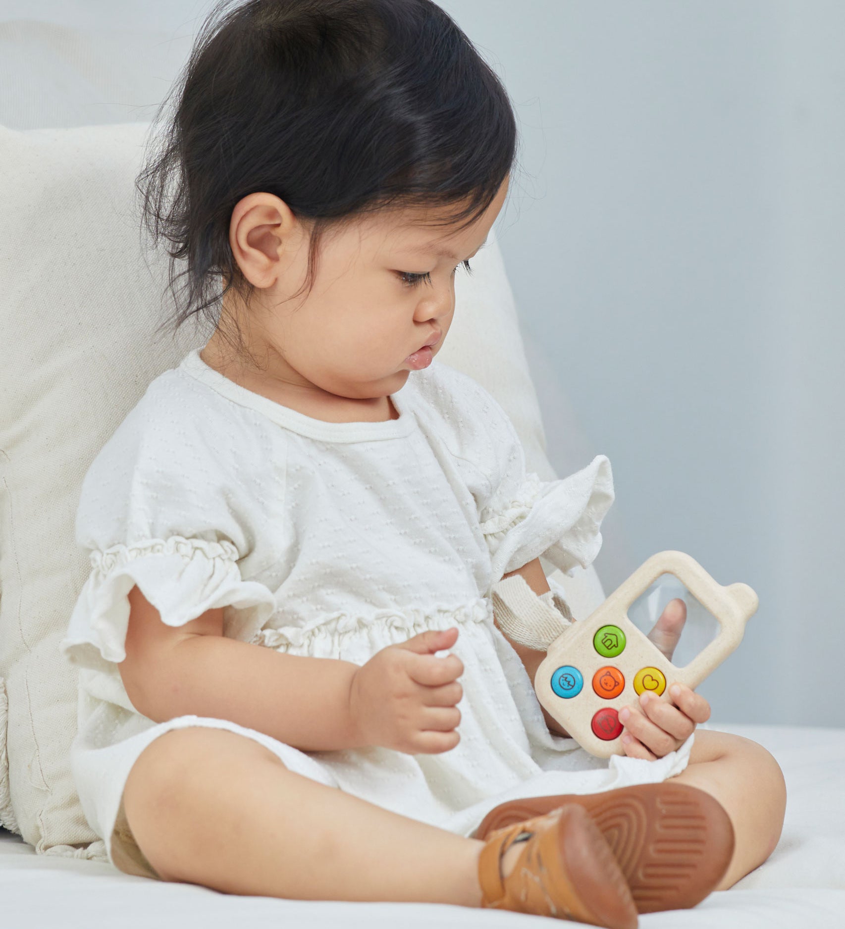 A baby sitting on a bed holding a PlanToys My First Phone toy. 