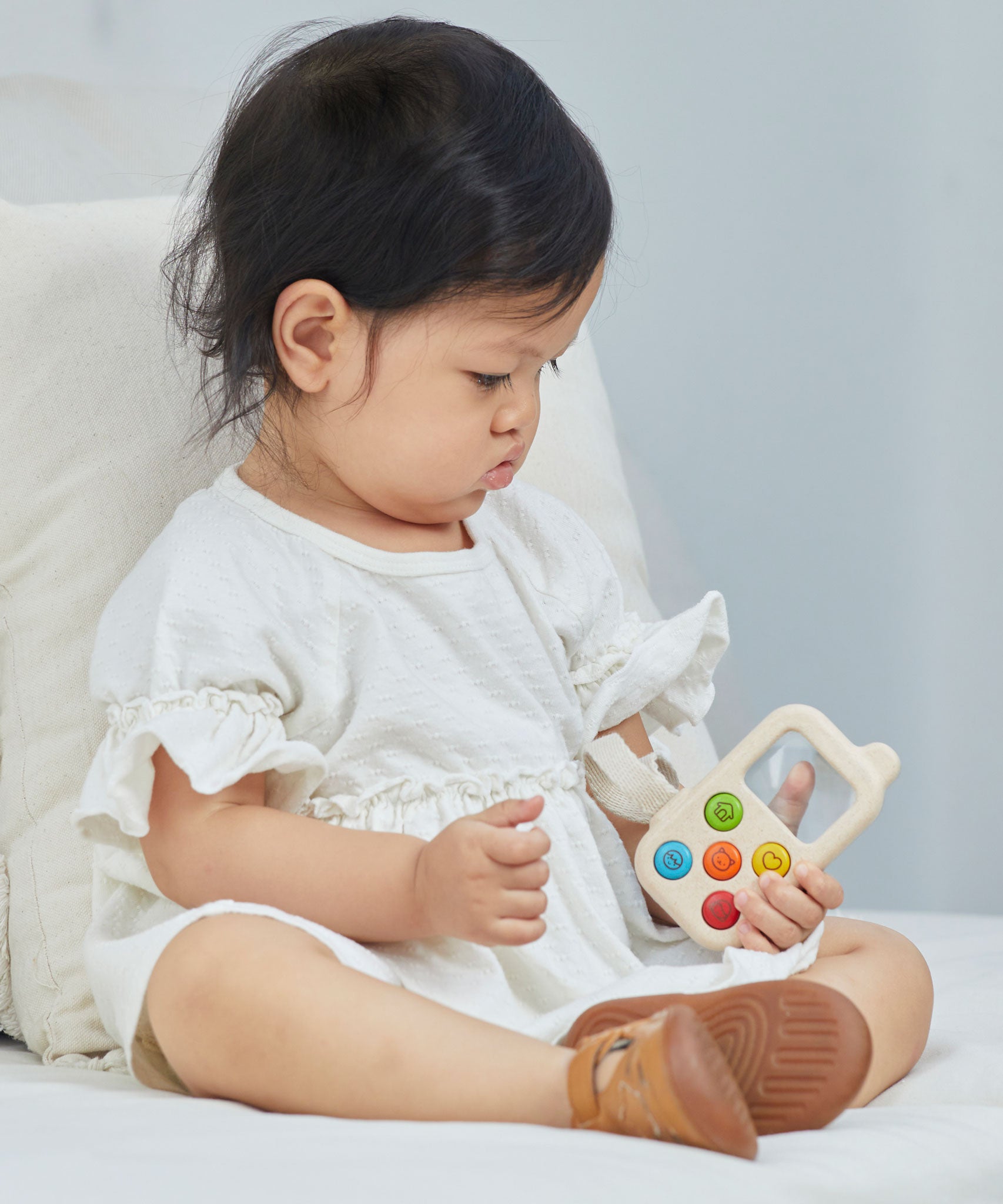 A baby sitting on a bed holding a PlanToys My First Phone toy. 