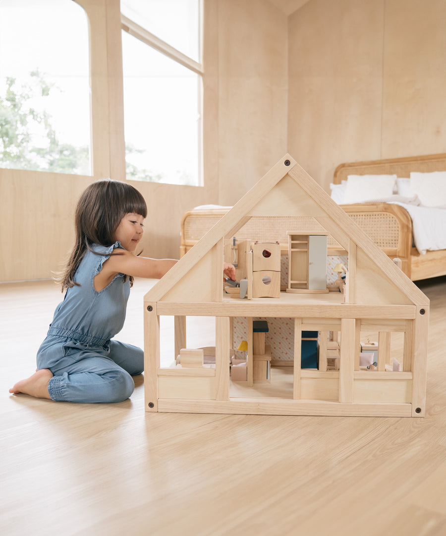 A young girl on her knees playing with the PlanToys eco-friendly dollhouse on a wooden bedroom floor.