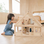 A young girl on her knees playing with the PlanToys eco-friendly dollhouse on a wooden bedroom floor.