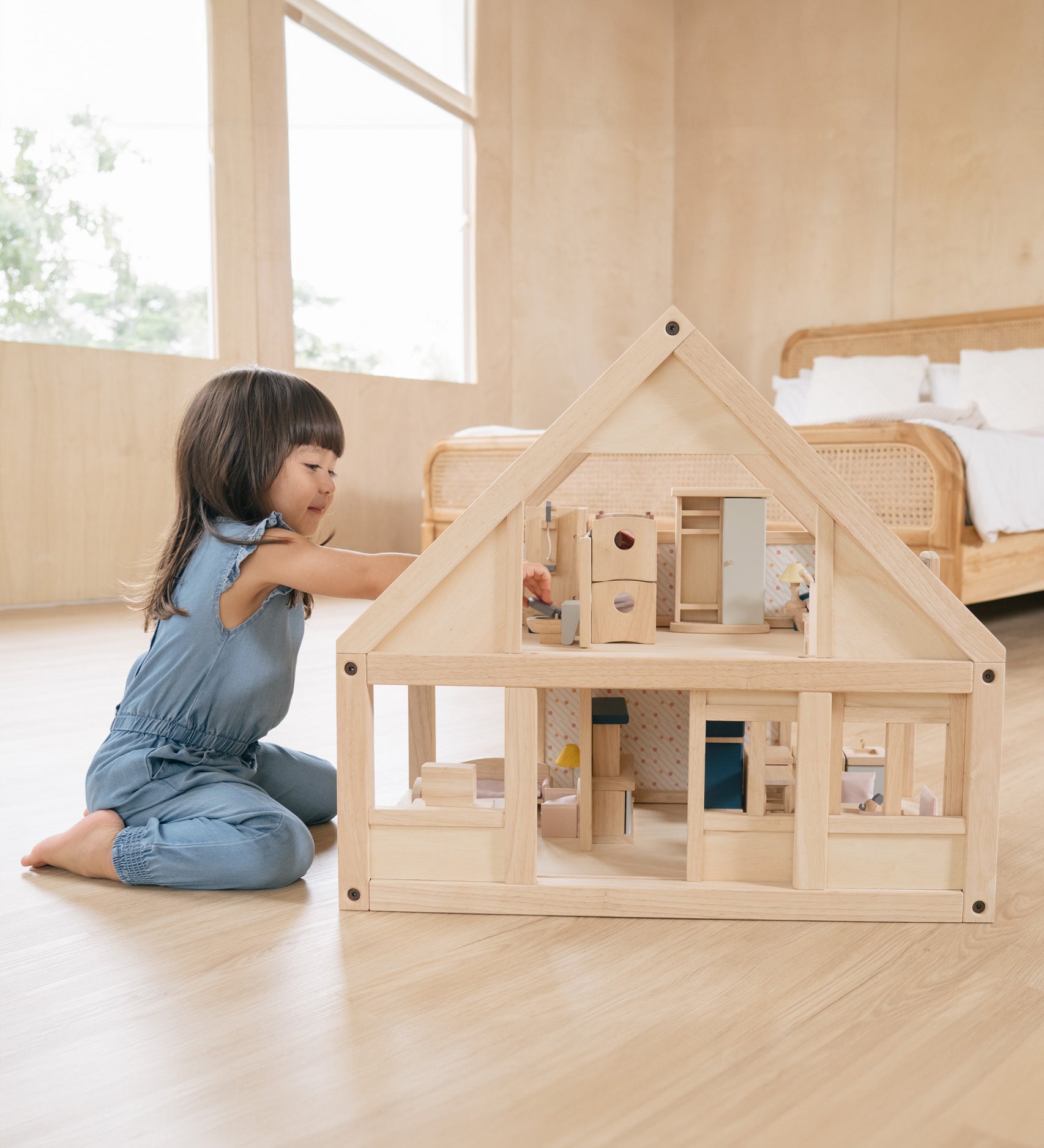 A young girl on her knees playing with the PlanToys eco-friendly dollhouse on a wooden bedroom floor.