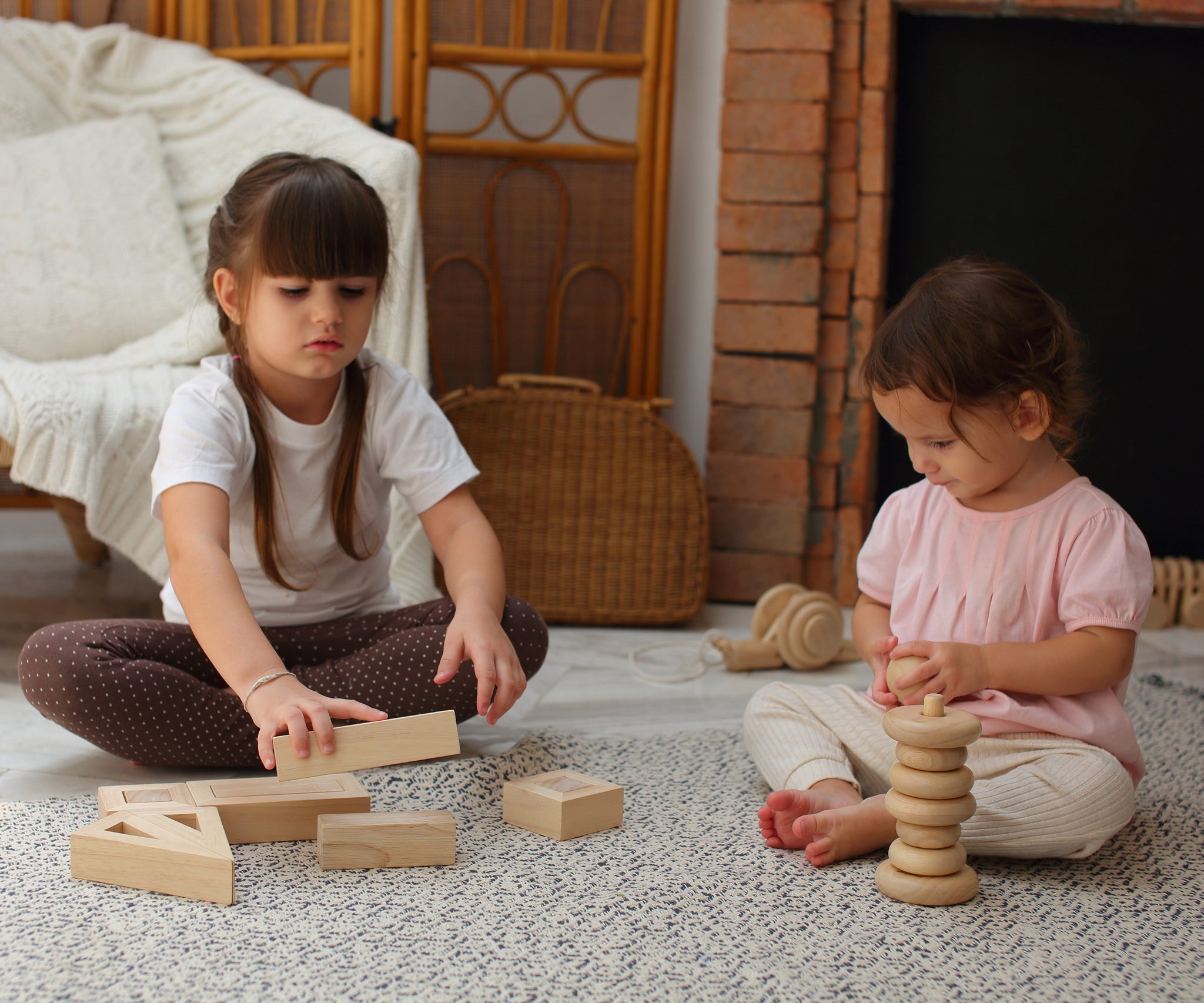 Two children sitting down on a patterned rug, one is playing with the PlanToys Hollow Blocks whilst the other is using the natural PlanToys stacker 