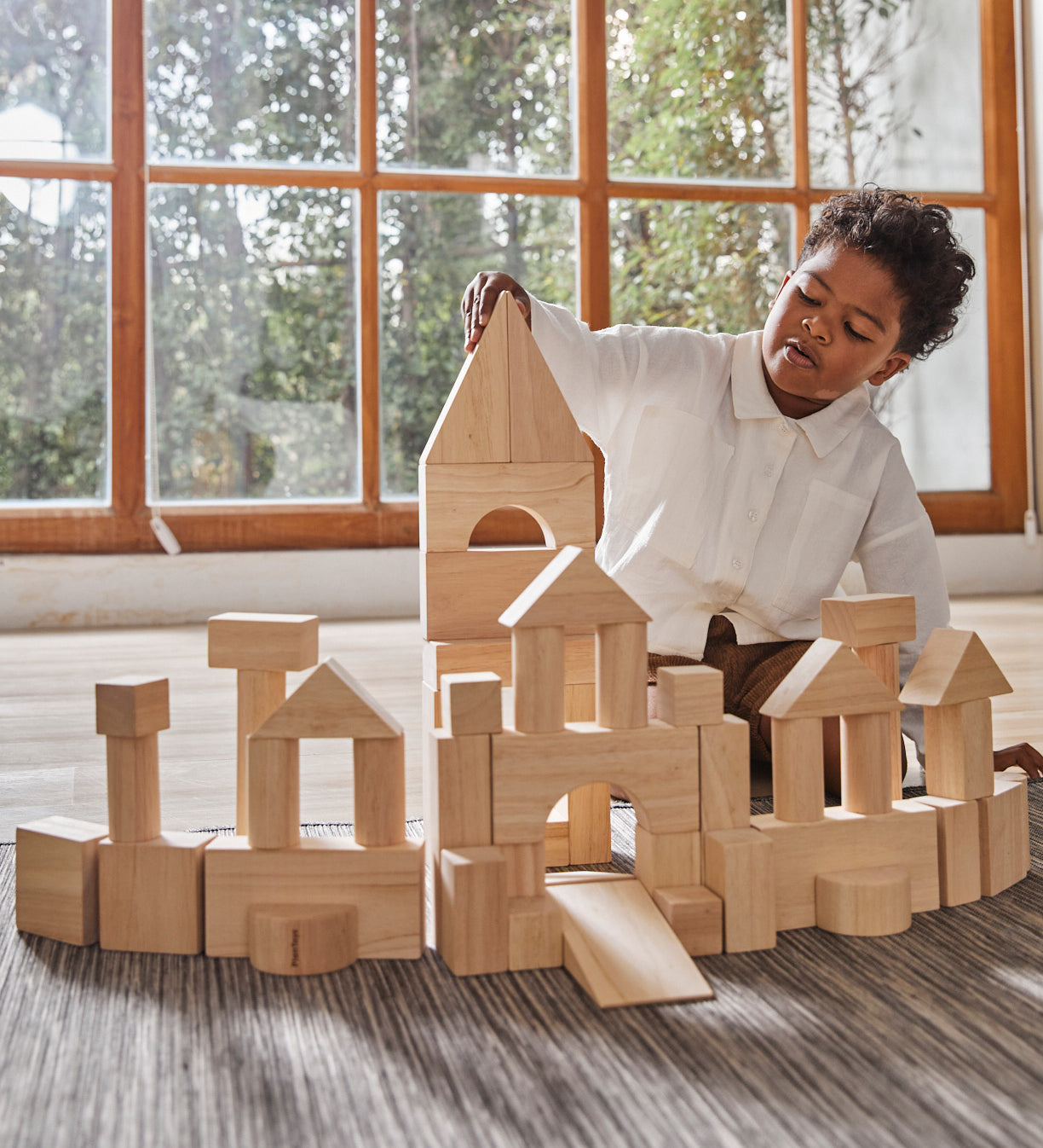 A child playing with the Plan Toys natural wooden 50 Unit Building Block set. The child has built a long building with multiple turret like pillars and a tall tower in the middle. The child can be seen sitting behind the structure. 
