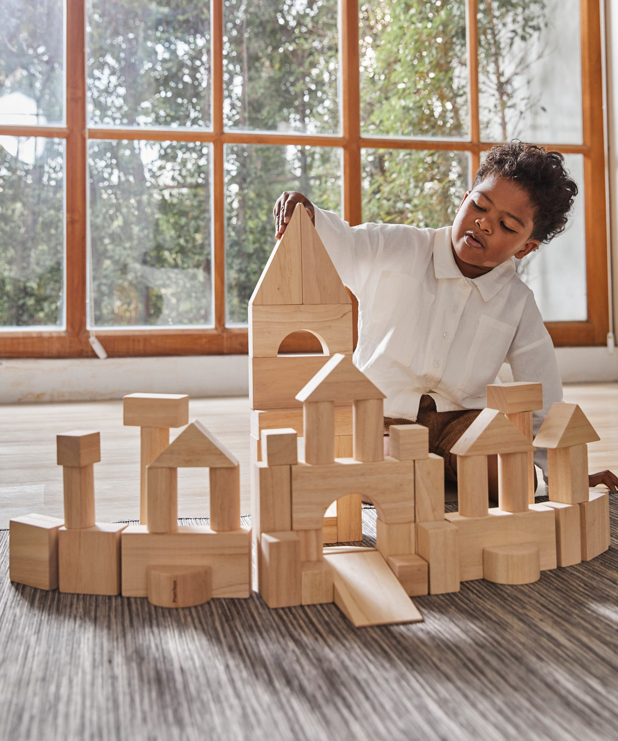 A child playing with the Plan Toys natural wooden 50 Unit Building Block set. The child has built a long building with multiple turret like pillars and a tall tower in the middle. The child can be seen sitting behind the structure. 
