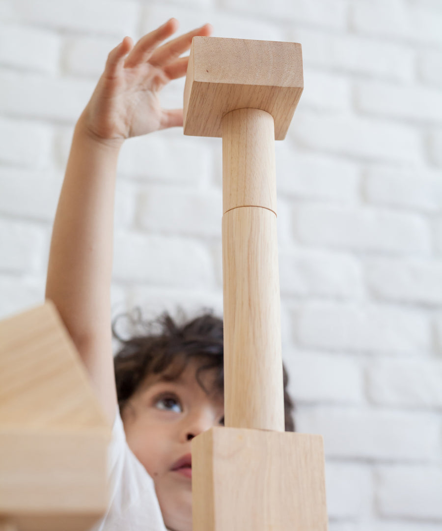 A close up of a child playing with the Plan Toys natural wooden 50 Unit Building Block set. the child can be seen looking up at a pile of clocks reaching for the square shaped block that has been stacked on top of cylinder shaped blocks. 