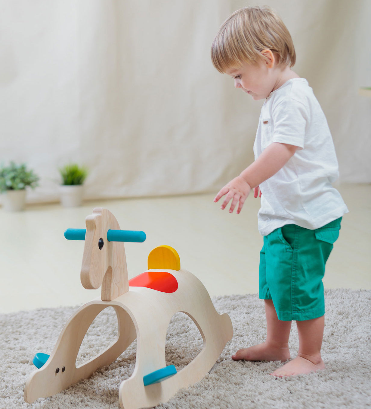 A child walking towards the PlanToys Palomino Rocking Horse. The rocking horse is on a grey coloured rug. 