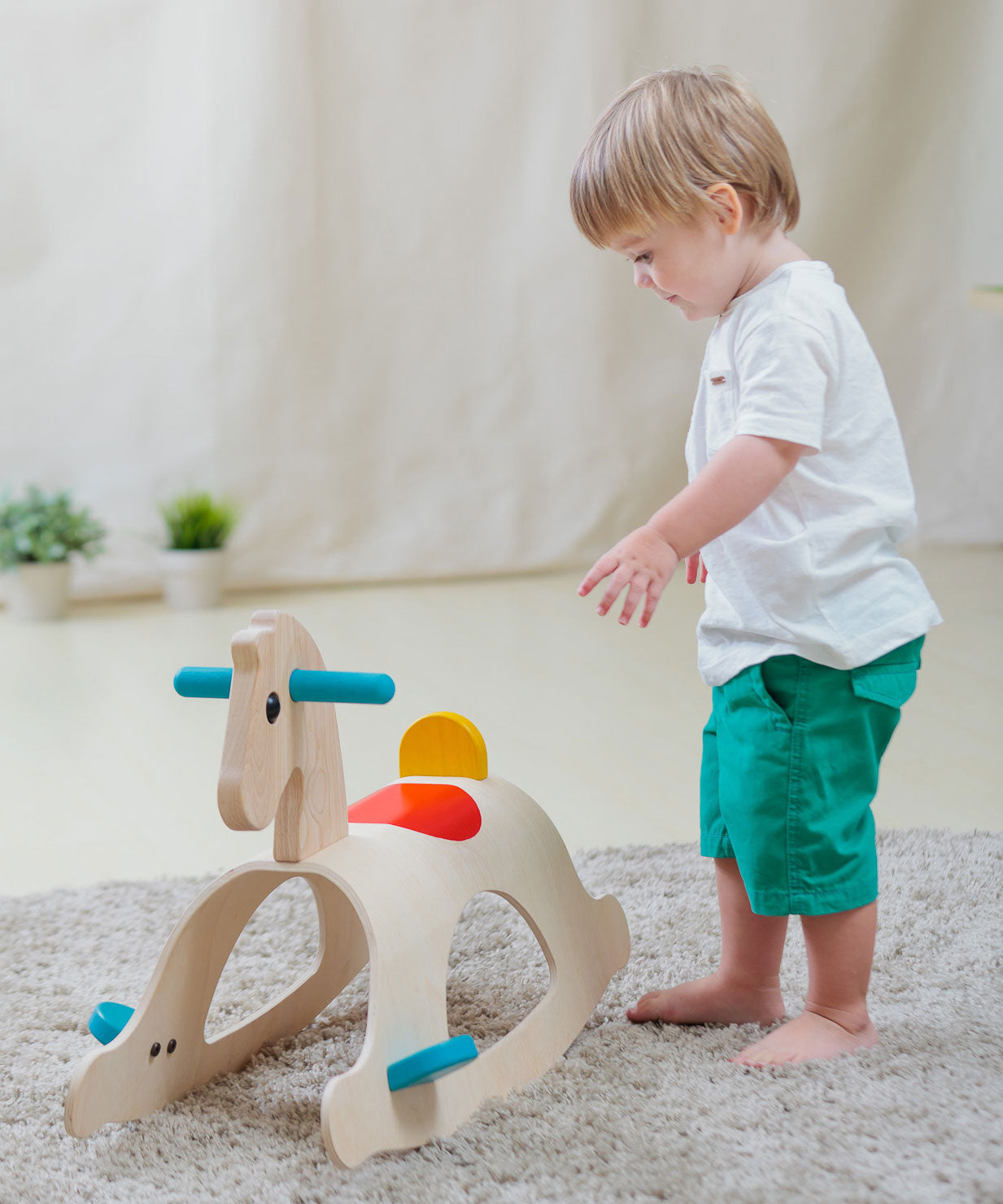 A child walking towards the PlanToys Palomino Rocking Horse. The rocking horse is on a grey coloured rug. 