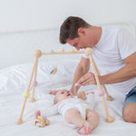 A PlanToys Pastel Play Gym placed on a bed, a baby is laying underneath looking up at an adult that is sat on the bed besides them 