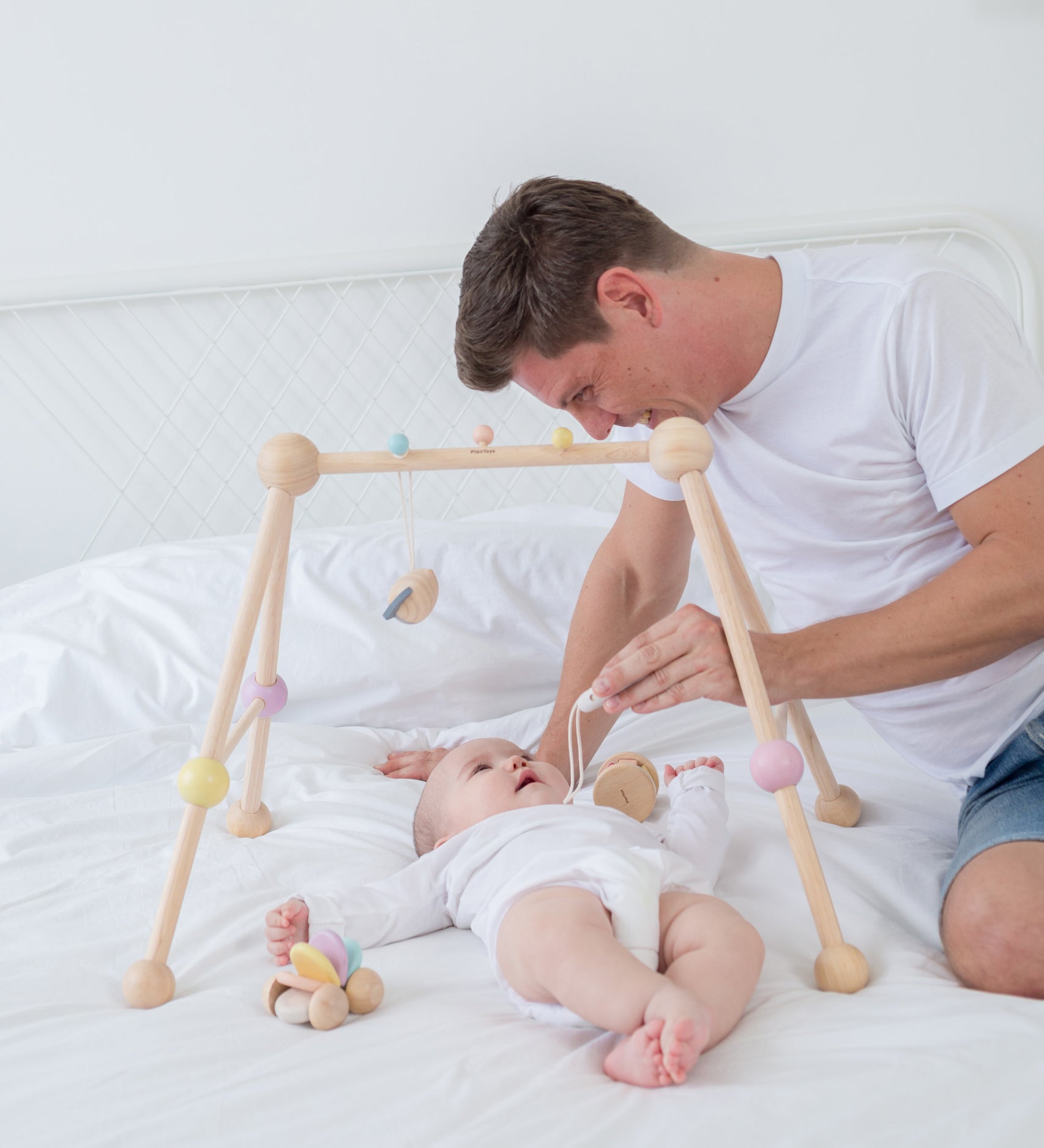 A PlanToys Pastel Play Gym placed on a bed, a baby is laying underneath looking up at an adult that is sat on the bed besides them 