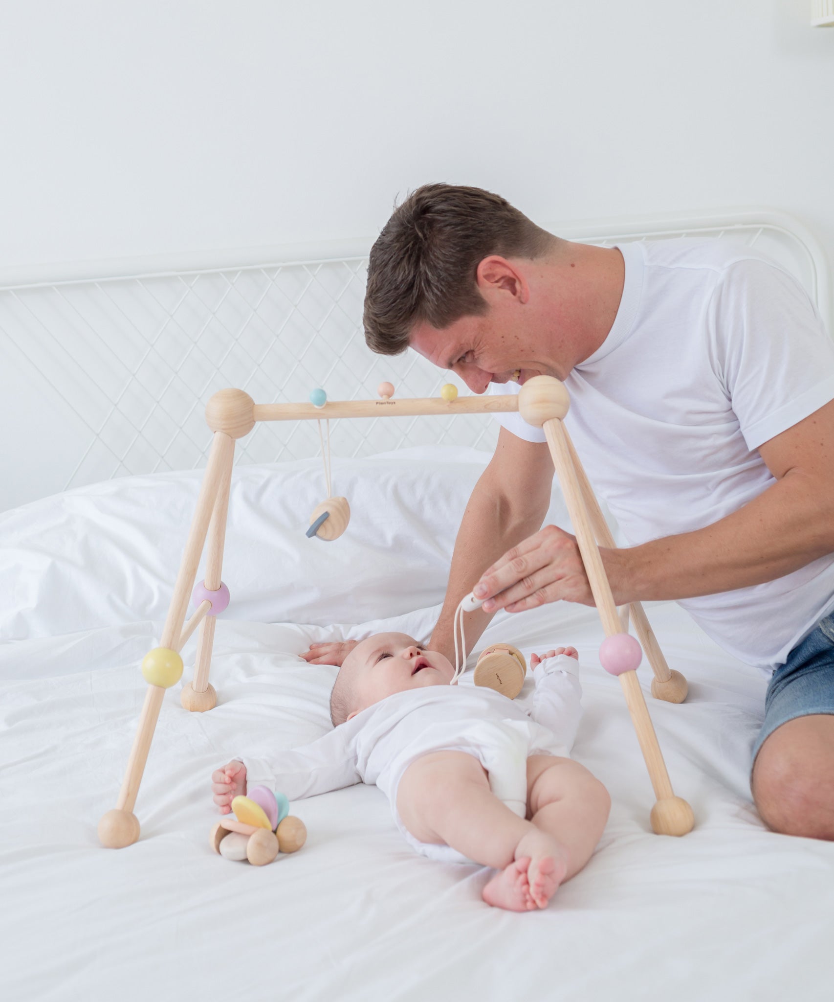 A PlanToys Pastel Play Gym placed on a bed, a baby is laying underneath looking up at an adult that is sat on the bed besides them 