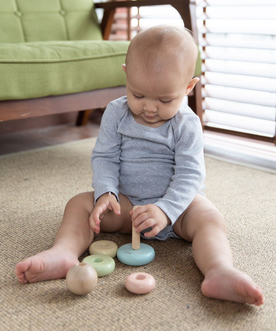 A baby playing with the PlanToys pastel baby stacking ring. The baby is sitting up on a jute style rug holding on to pole with the coloured rings all taken off. 