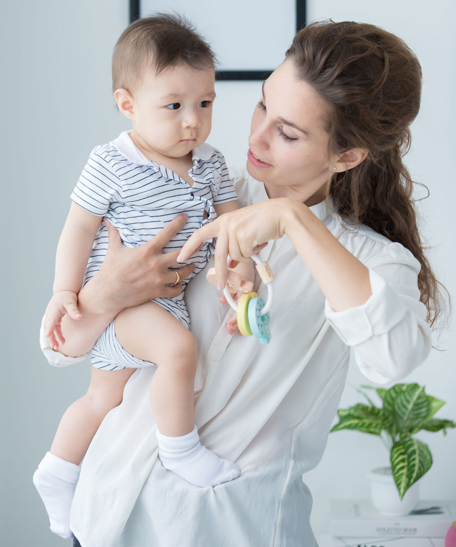 An adult holding a baby in one arm and a PlanToys Pastel Baby Key Rattle in the other hand. 
