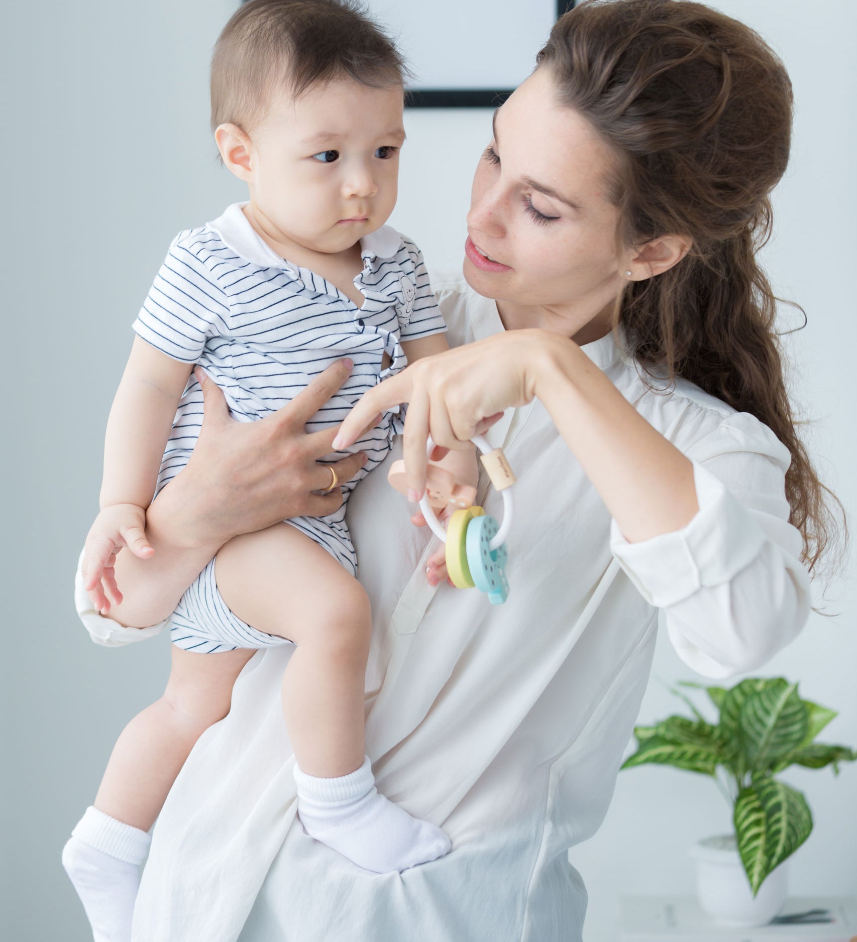 An adult holding a baby in one arm and a PlanToys Pastel Baby Key Rattle in the other hand. 
