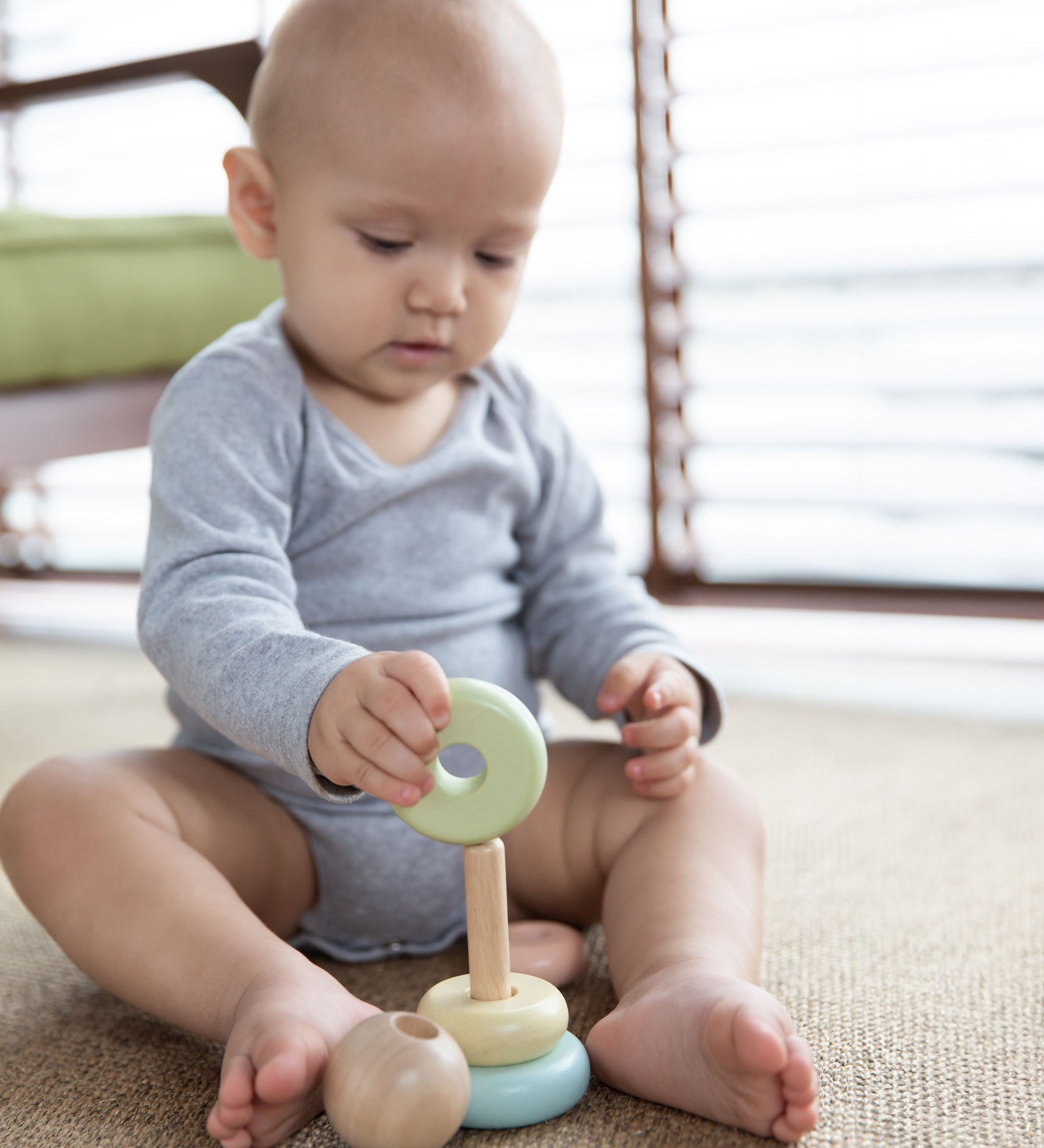 A baby playing with the PlanToys pastel baby stacking ring. The baby is sitting up on a jute style rug holding on to the green coloured ring. 