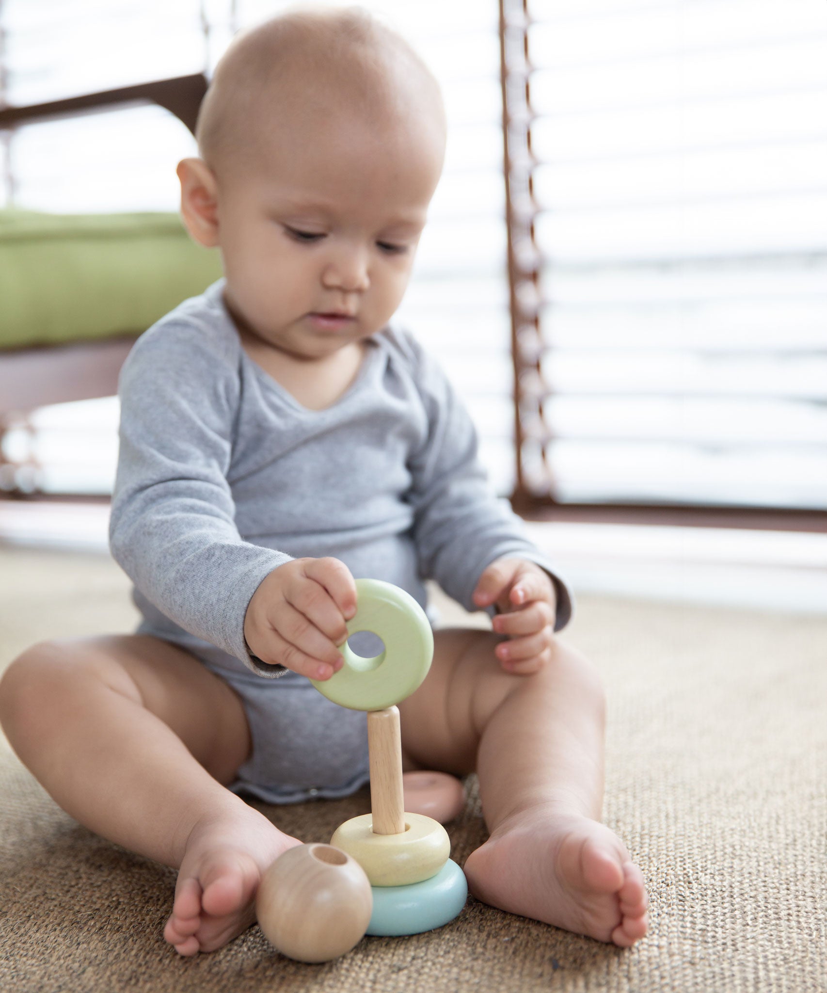 A baby playing with the PlanToys pastel baby stacking ring. The baby is sitting up on a jute style rug holding on to the green coloured ring. 