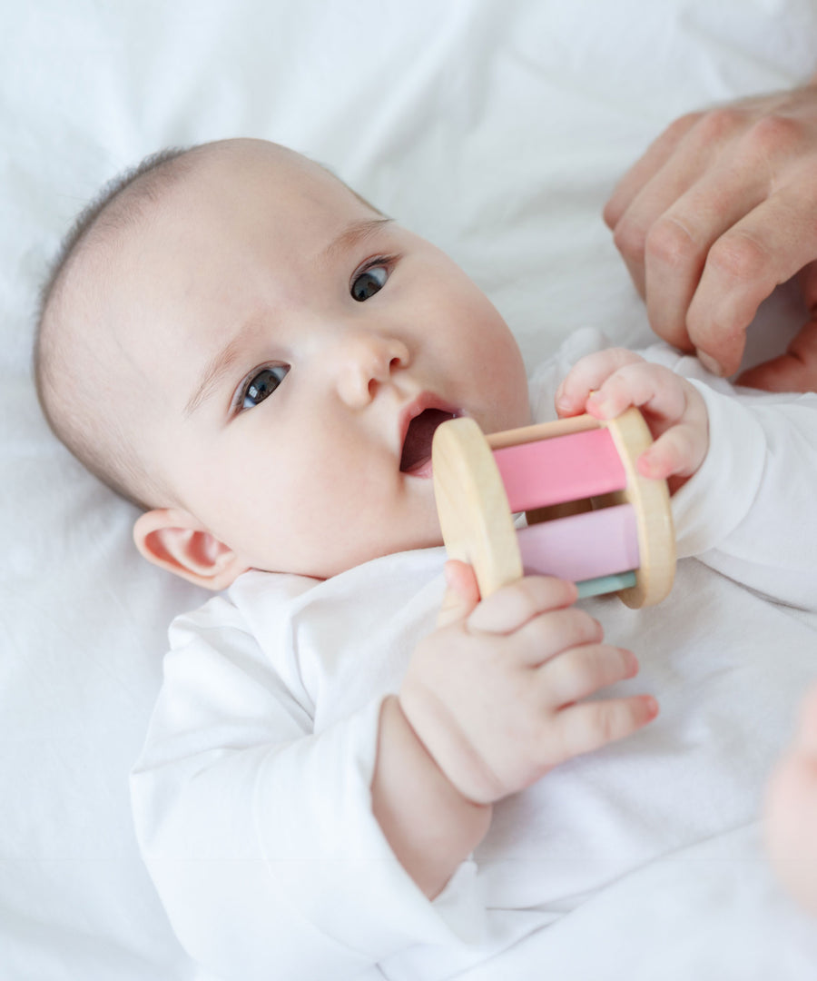 A baby is laying down on a white sheet holding a PlanToys Pastel Roller up to their mouth. 