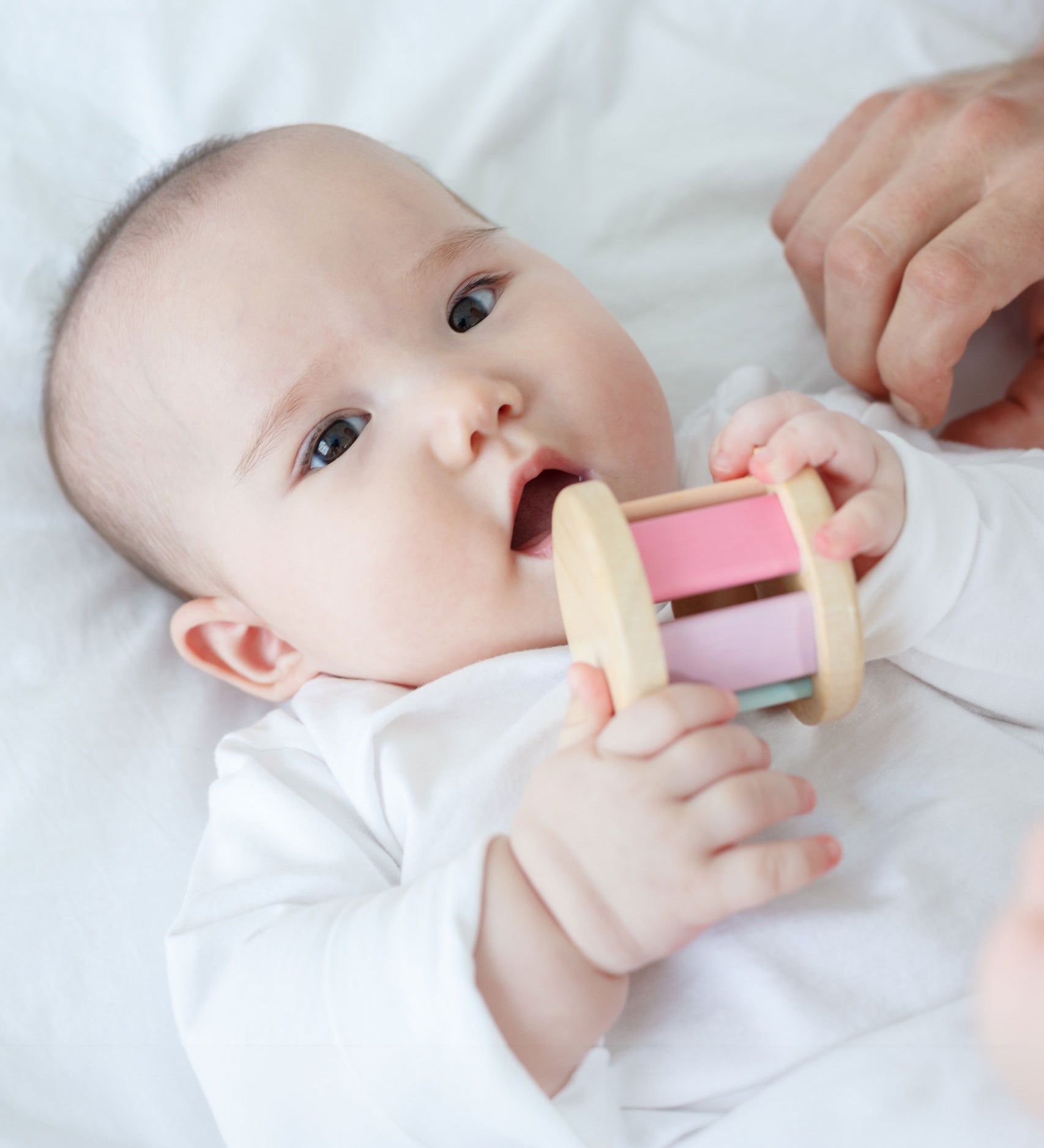 A baby is laying down on a white sheet holding a PlanToys Pastel Roller up to their mouth. 