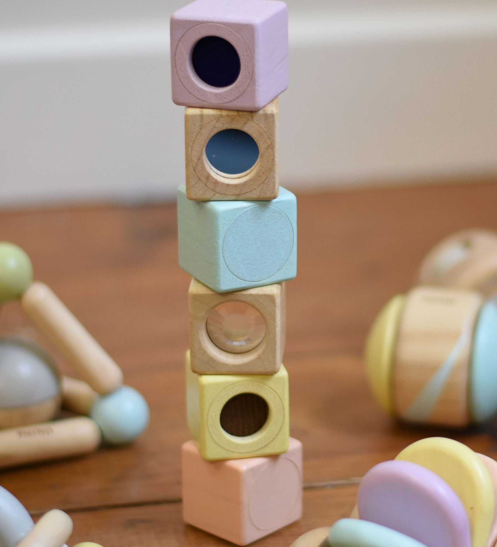 The PlanToys Pastel Sensory Blocks stacked up on a wooden floor. Other PlanToys pastel baby toys can be seen around them.