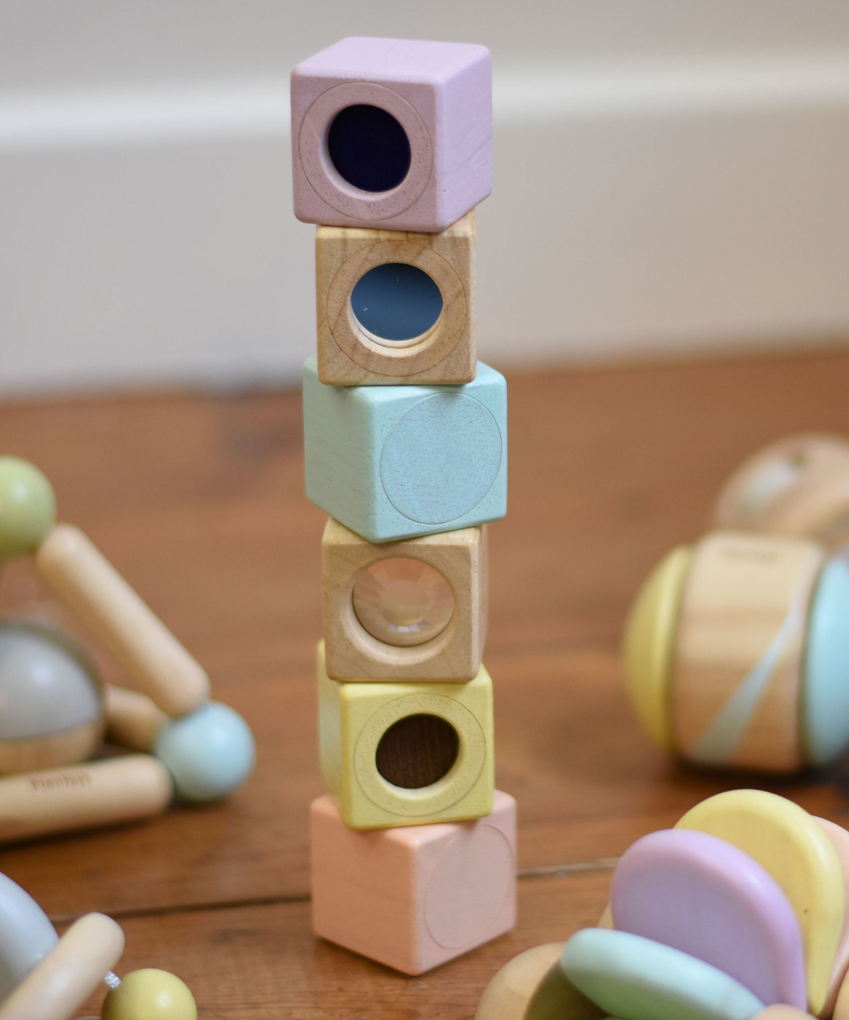 The PlanToys Pastel Sensory Blocks stacked up on a wooden floor. Other PlanToys pastel baby toys can be seen around them.