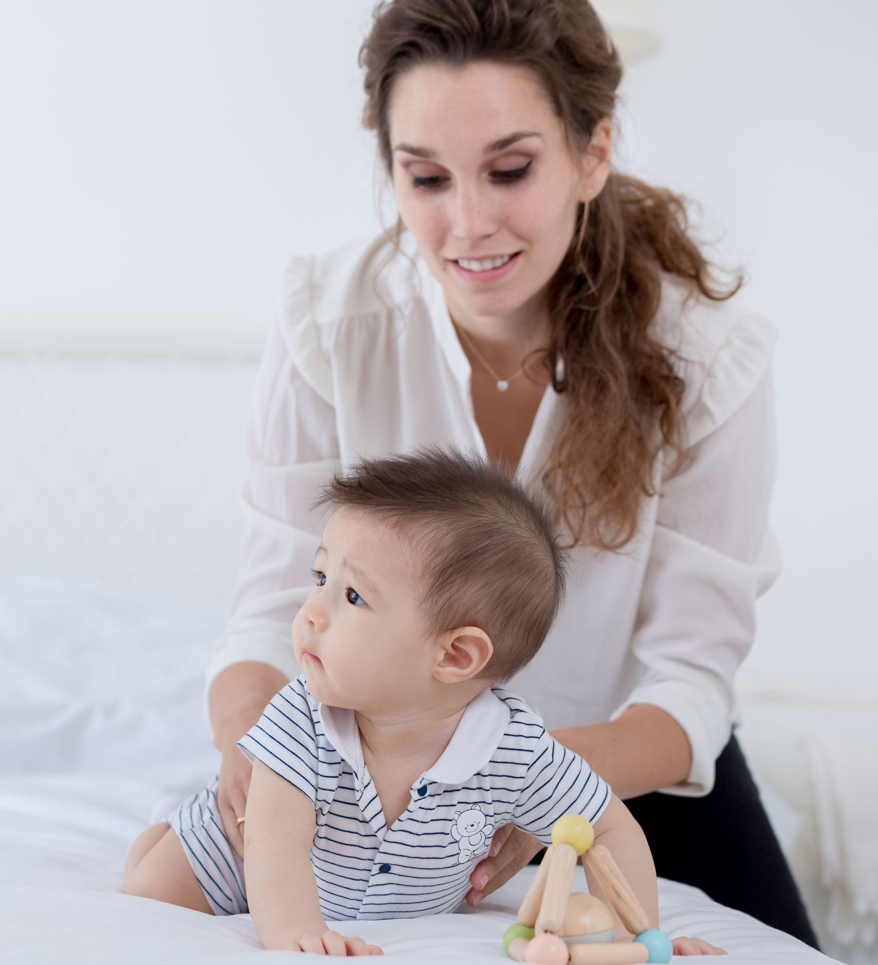 A bay playing with a PlanToys Pastel Triangle Clutching Toy on a white bed, an adult is holding onto them. 