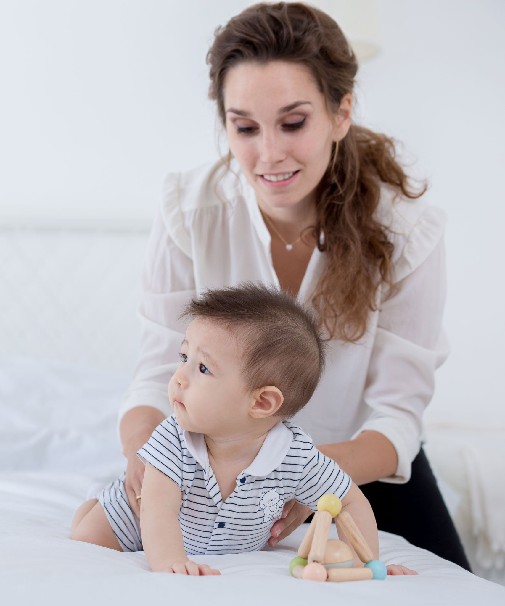 A bay playing with a PlanToys Pastel Triangle Clutching Toy on a white bed, an adult is holding onto them. 