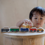 The PlanToys Planworld Cars all lined up on a wooden stool, a child can be seen standing behind them reaching for the red car. 