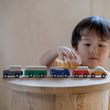 The PlanToys Planworld Cars all lined up on a wooden stool, a child can be seen standing behind them reaching for the red car. 