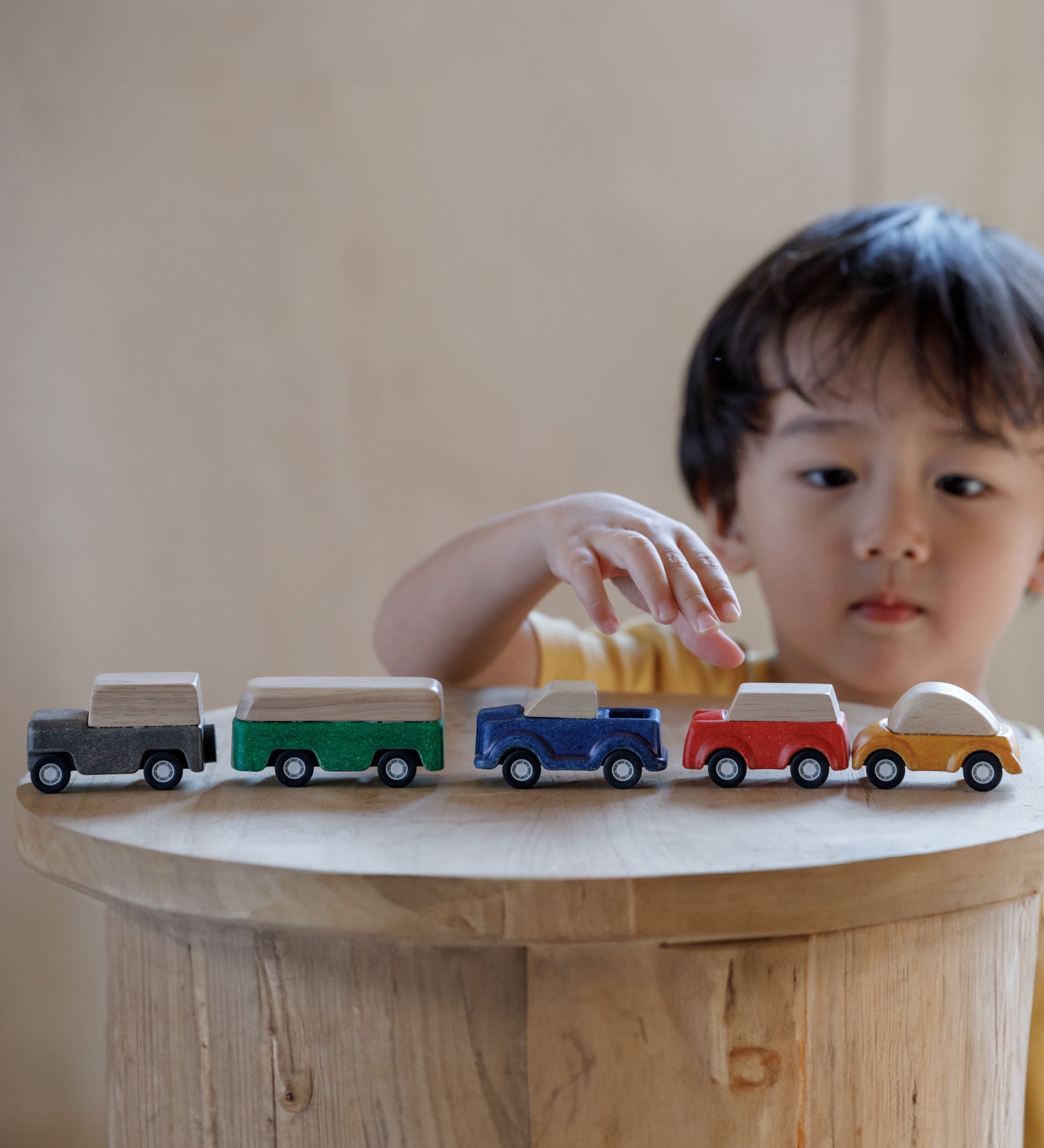 The PlanToys Planworld Cars all lined up on a wooden stool, a child can be seen standing behind them reaching for the red car. 