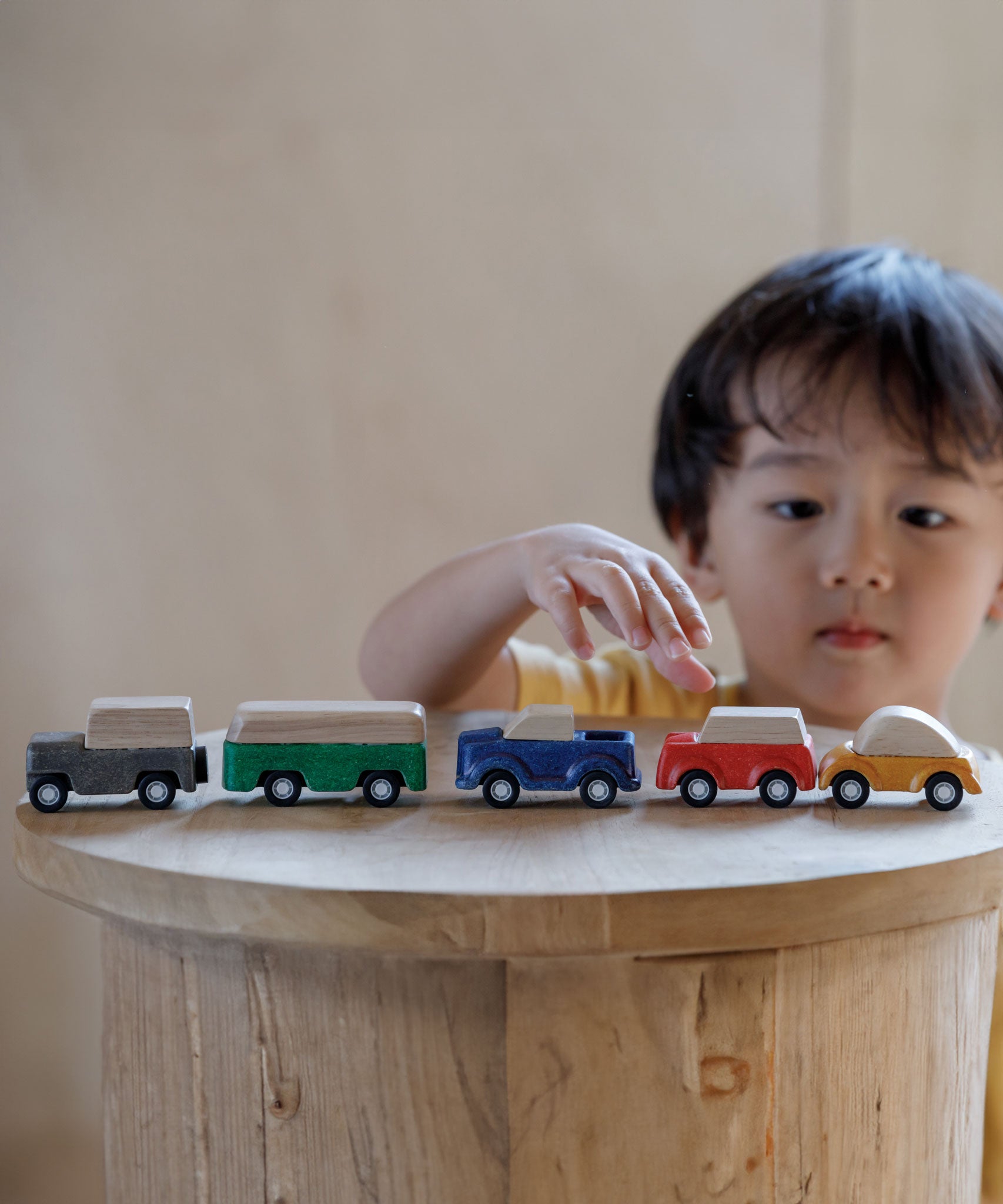 The PlanToys Planworld Cars all lined up on a wooden stool, a child can be seen standing behind them reaching for the red car. 