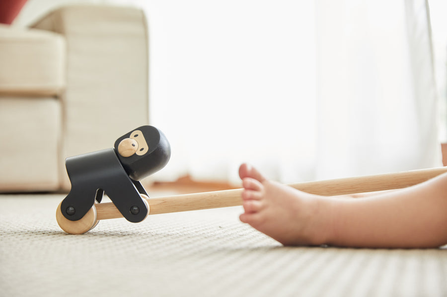 Close up of the PlanToys wooden gorilla pull along toy on a white carpet next to a child's feet