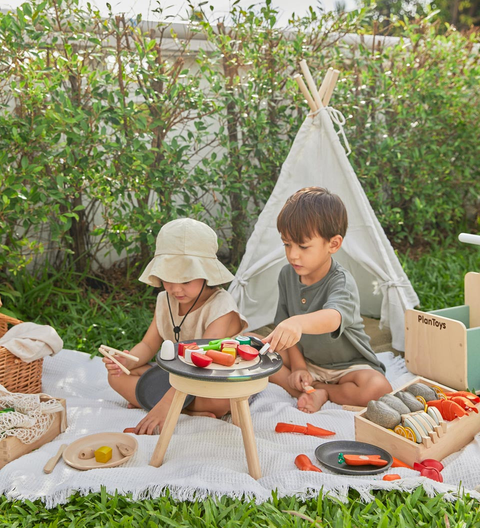 Two children sat on a blanket in front of a small tent playing with wooden play food on top of the PlanToys wooden BBQ set