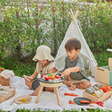 Two children sat on a blanket in front of a small tent playing with wooden play food on top of the PlanToys wooden BBQ set