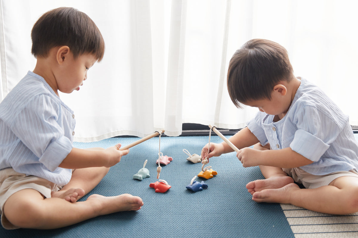 Two children sat on a blue carpet playing with the PlanToys fish hooking game