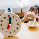 Young child crouched down holding a teddy bear next to an empty bowl beside the PlanToys wooden activity clock