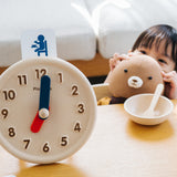 Young child crouched down holding a teddy bear next to an empty bowl beside the PlanToys wooden activity clock