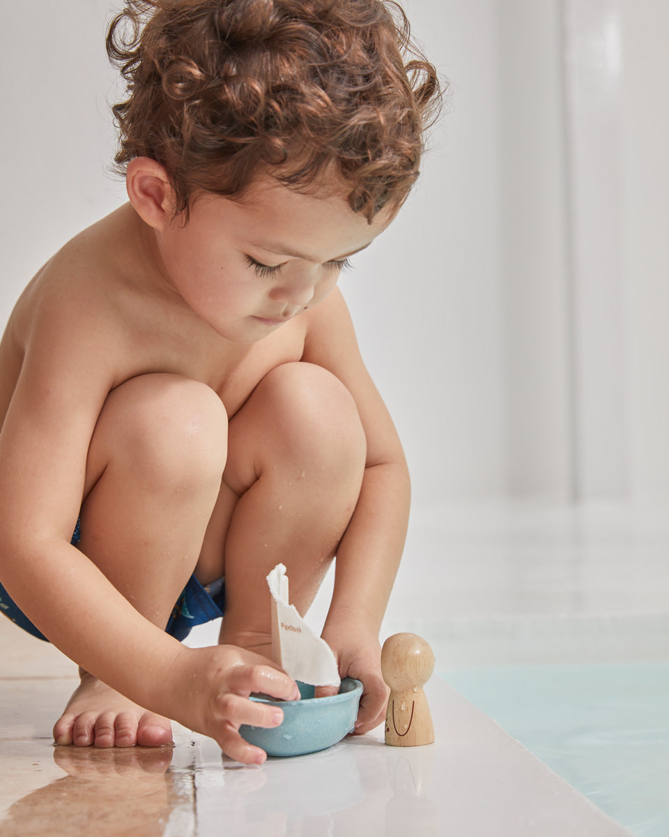 Close up of young boy crouched down playing with the PlanToys wooden walrus sailing boat toy next to a swimming pool