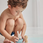 Close up of young boy crouched down playing with the PlanToys wooden walrus sailing boat toy next to a swimming pool