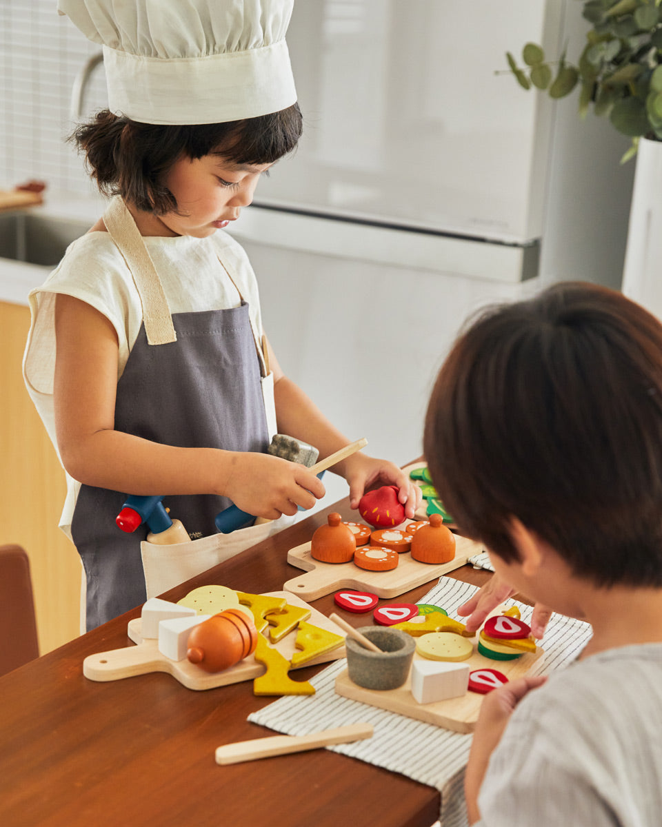 Young girl dressed as a chef playing with the PlanToys wooden play food cheese and charcuterie board set on a wooden worktop