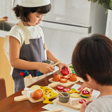 Young girl dressed as a chef playing with the PlanToys wooden play food cheese and charcuterie board set on a wooden worktop